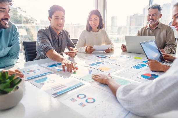 Multi racial diverse group of people working with Paperwork on a board room table at a business presentation or seminar. The documents have financial or marketing figures, graphs and charts on them. People are pointing to different documents. There are laptops and digital tablets on the table